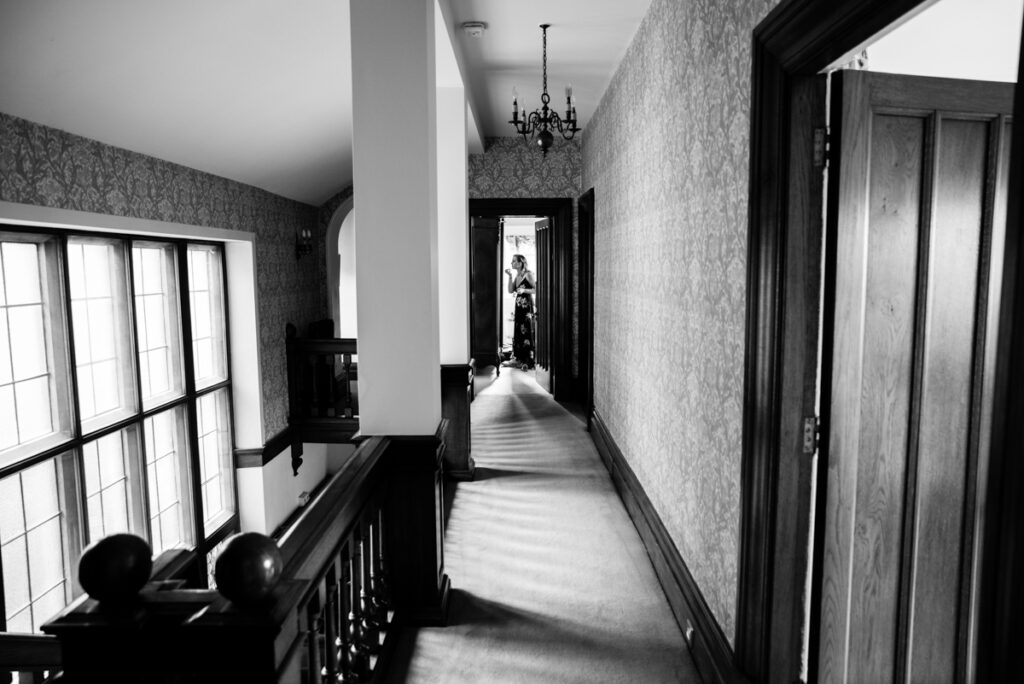 Black and white documentary photograph of a bridesmaid at the end of a corridor in an elegant house