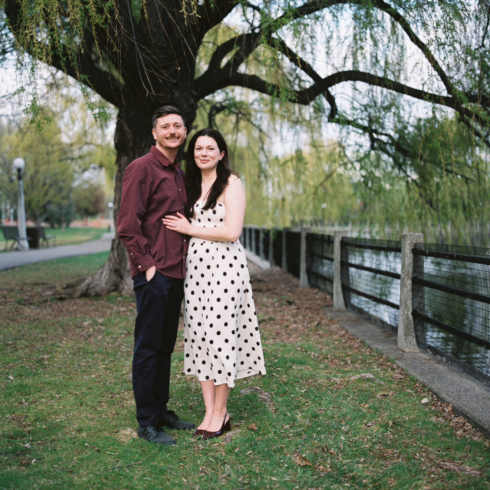 Ottawa Wedding Photographer Sara-0012 ❥ Ottawa Wedding Photographer Smiling couple stands close together on a grassy park path under a large tree near a railing by the water. Film Photography Bronica Zenza